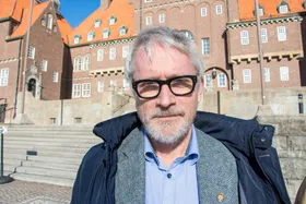 Anders Kumlin, en man med grått hår, skägg och glasögon, framför en byggnad med rött tak i solljus.