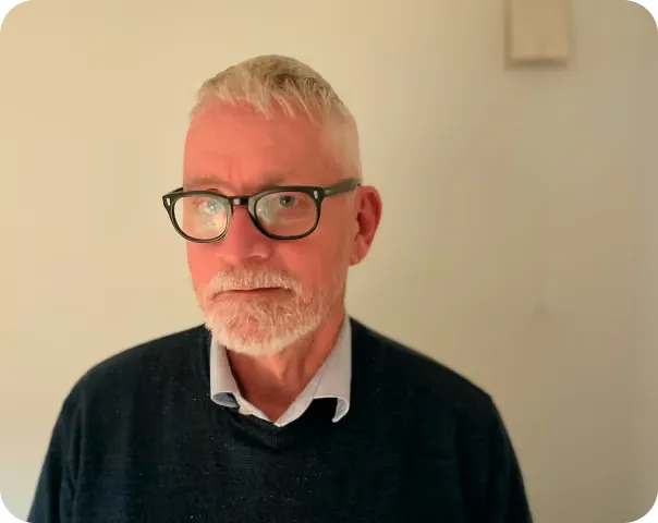 Anders Kumlin, en äldre man med glasögon, grått hår och skägg, i mörk tröja och ljus skjorta.